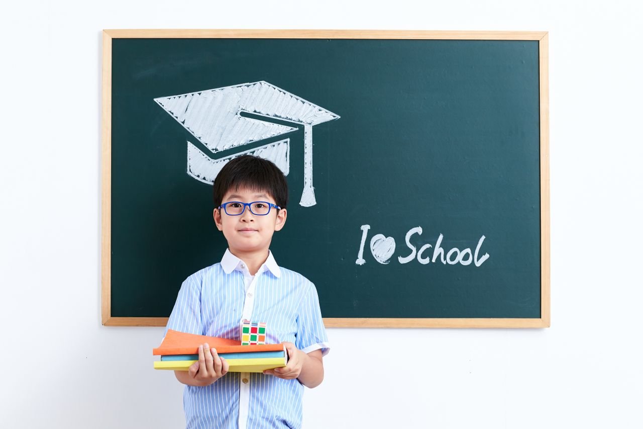 A smiling young boy stands in front of a chalkboard with a graduation cap drawing and "I love school" text.